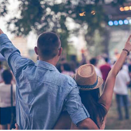 Personas disfrutando de un evento al aire libre, con los brazos levantados frente a un escenario y actividades de entretenimiento en vivo.
