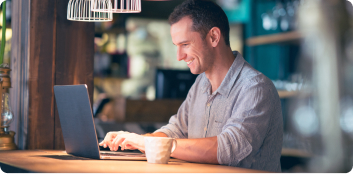 Un hombre sonriendo mientras usa una laptop y toma un café.