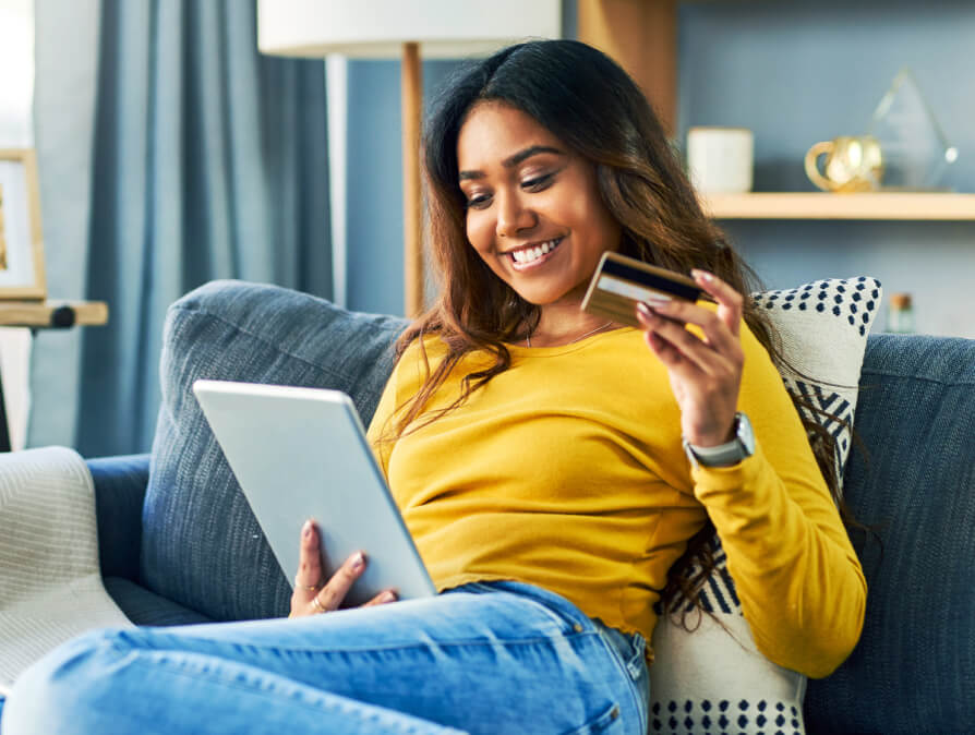 Una chica sentada en su sillón revisa su tablet mientras sostiene su tarjeta de débito.